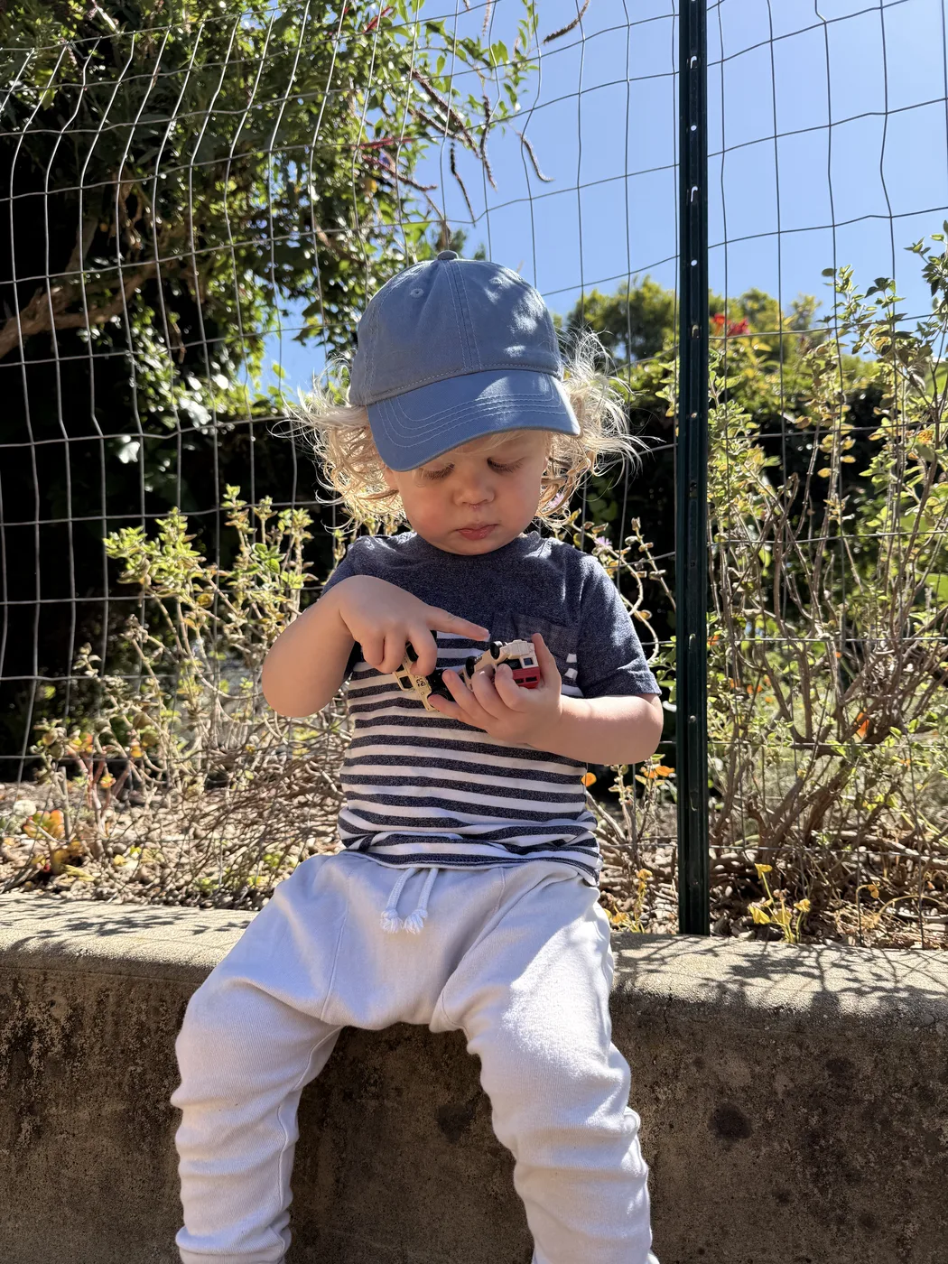 A toddler in a sun hat sitting on a low garden wall inspecting a small toy
