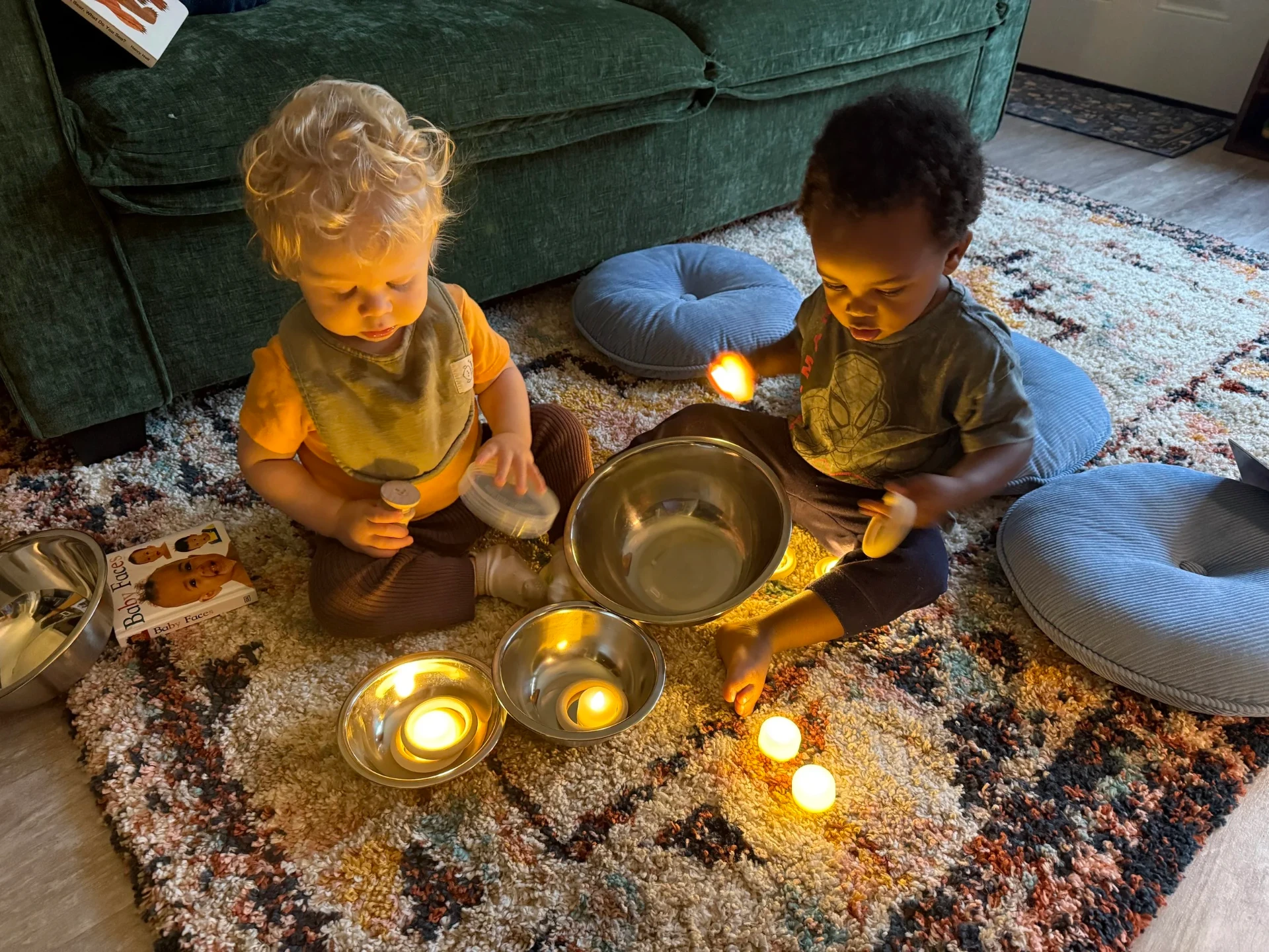 Two toddlers exploring warm candlelight and small metal bowls together on the rug