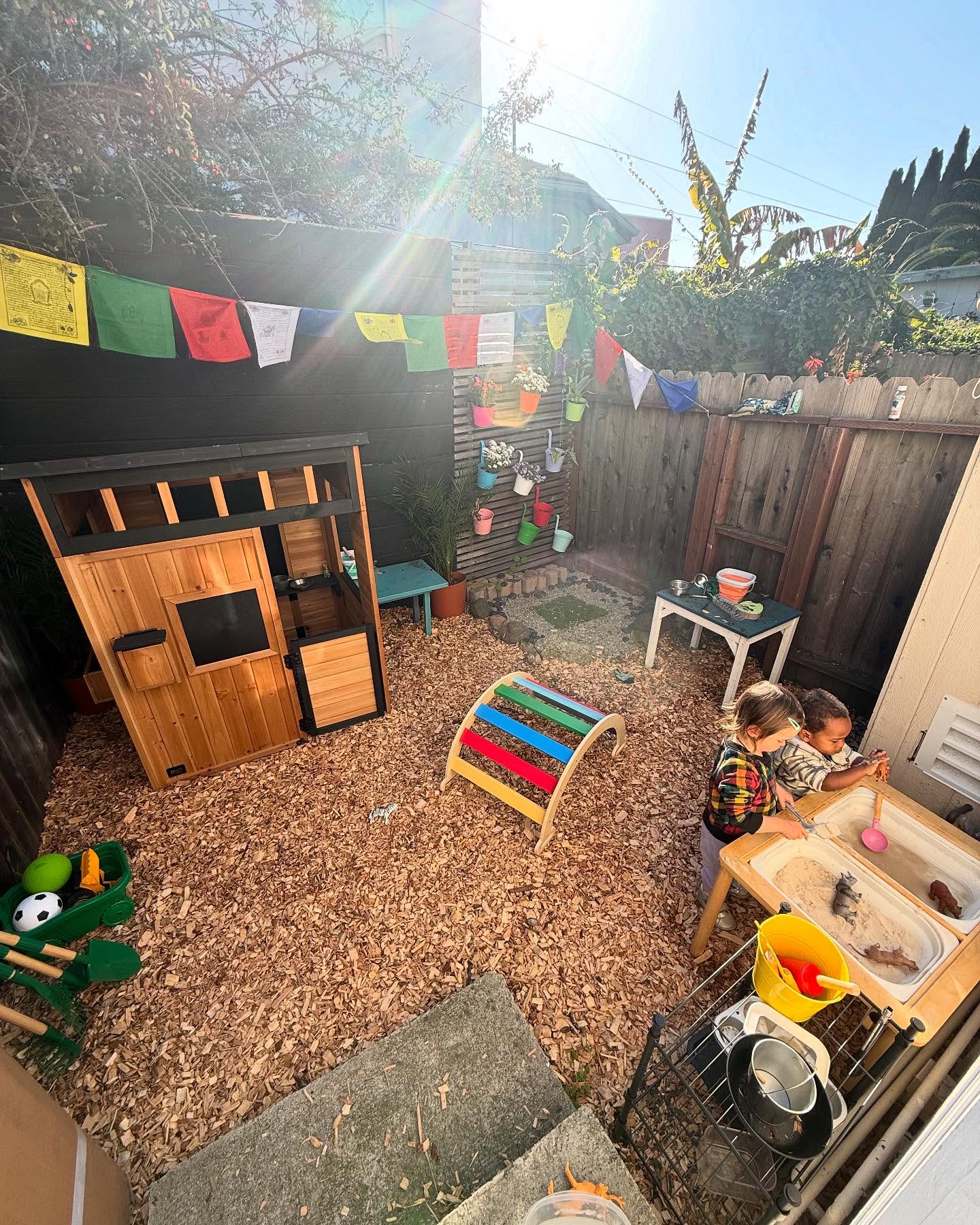 Sun-drenched backyard play area with a wooden playhouse and toddlers at the sensory table