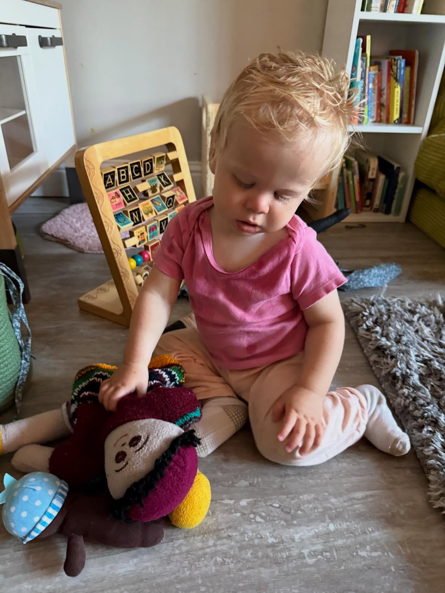 A toddler sitting on the floor with a soft doll and an alphabet board