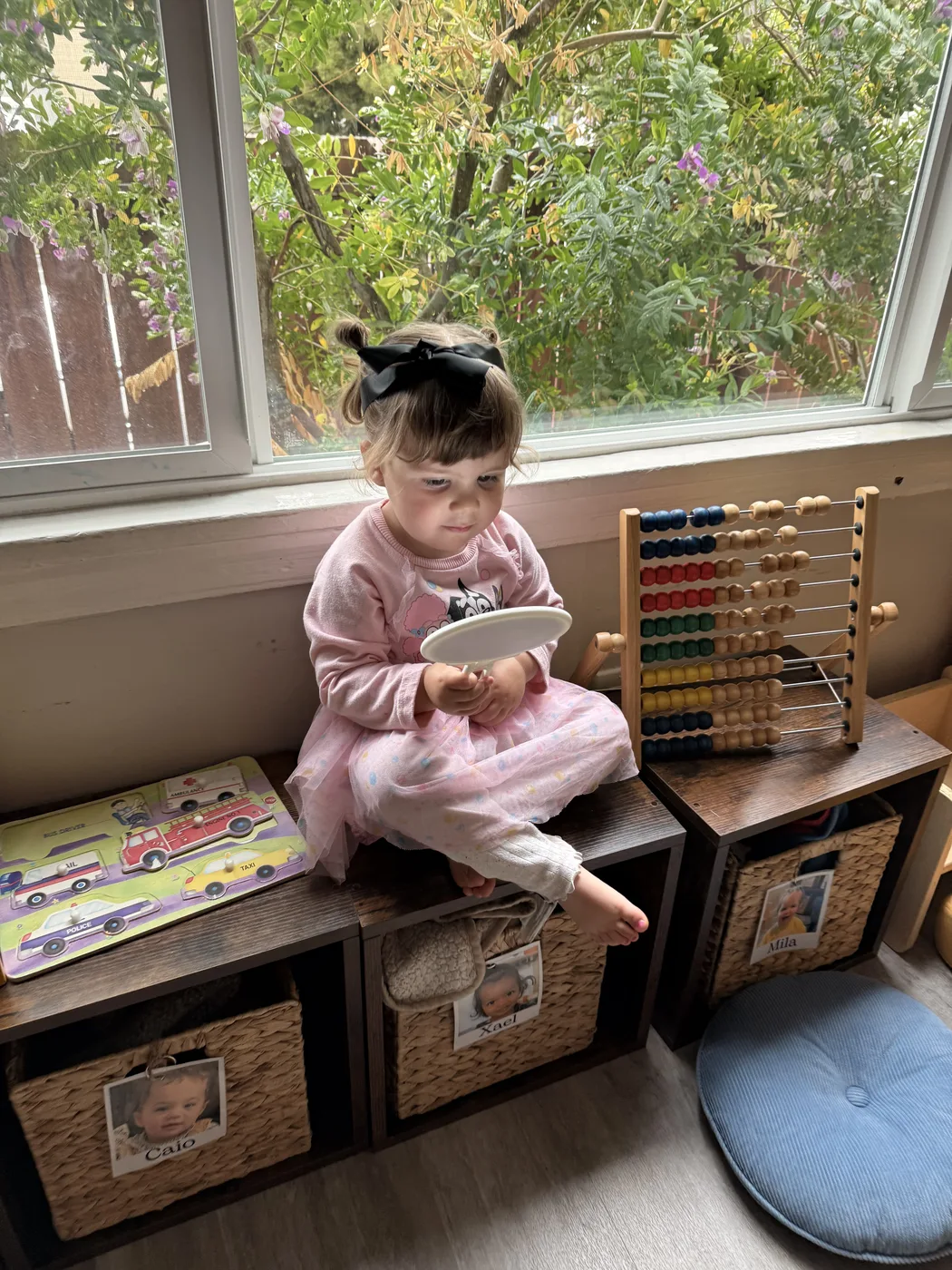 A toddler sitting by the window beside an abacus and a basket of books