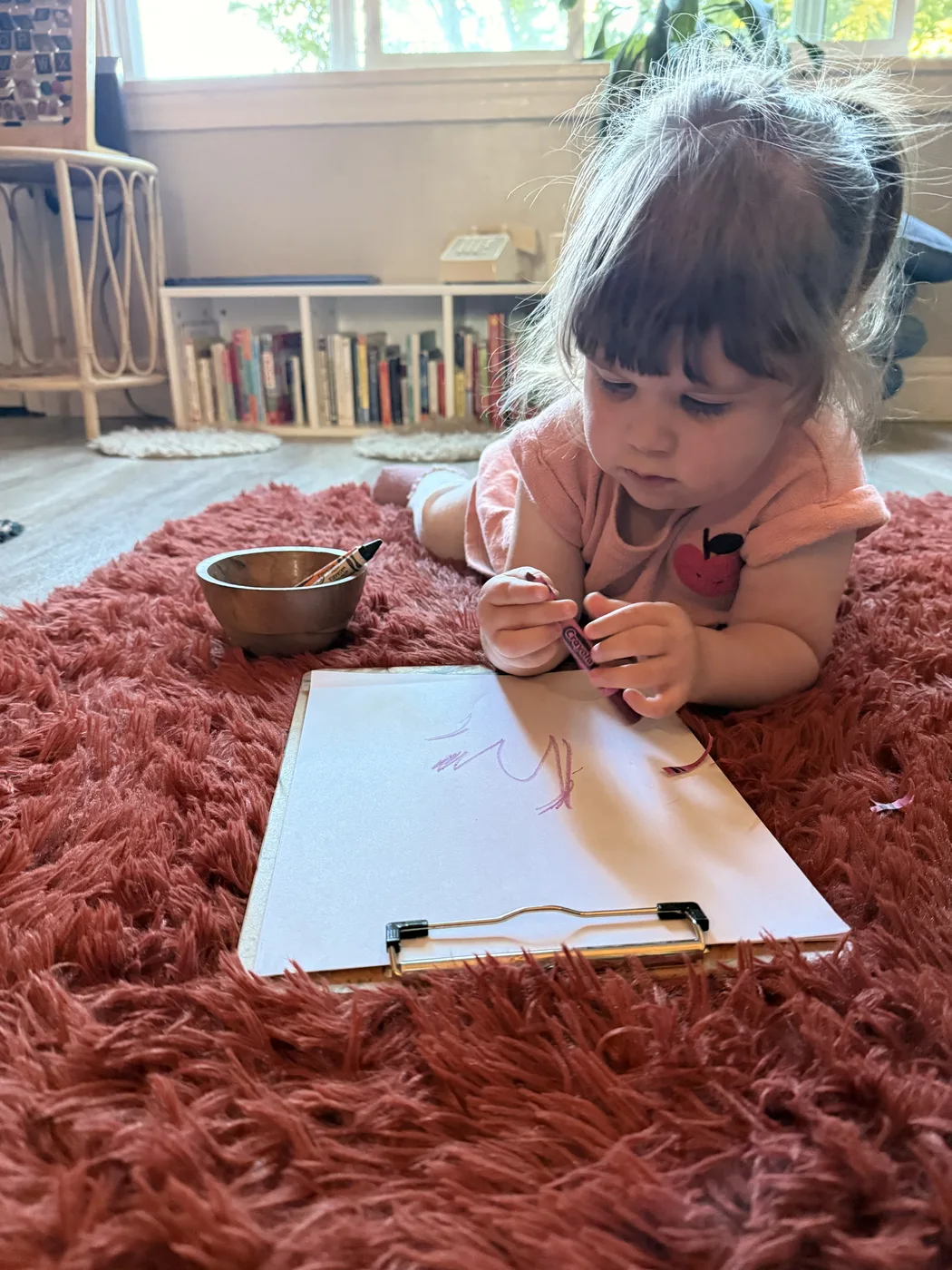 A toddler drawing with a crayon on paper resting on a rust-colored rug