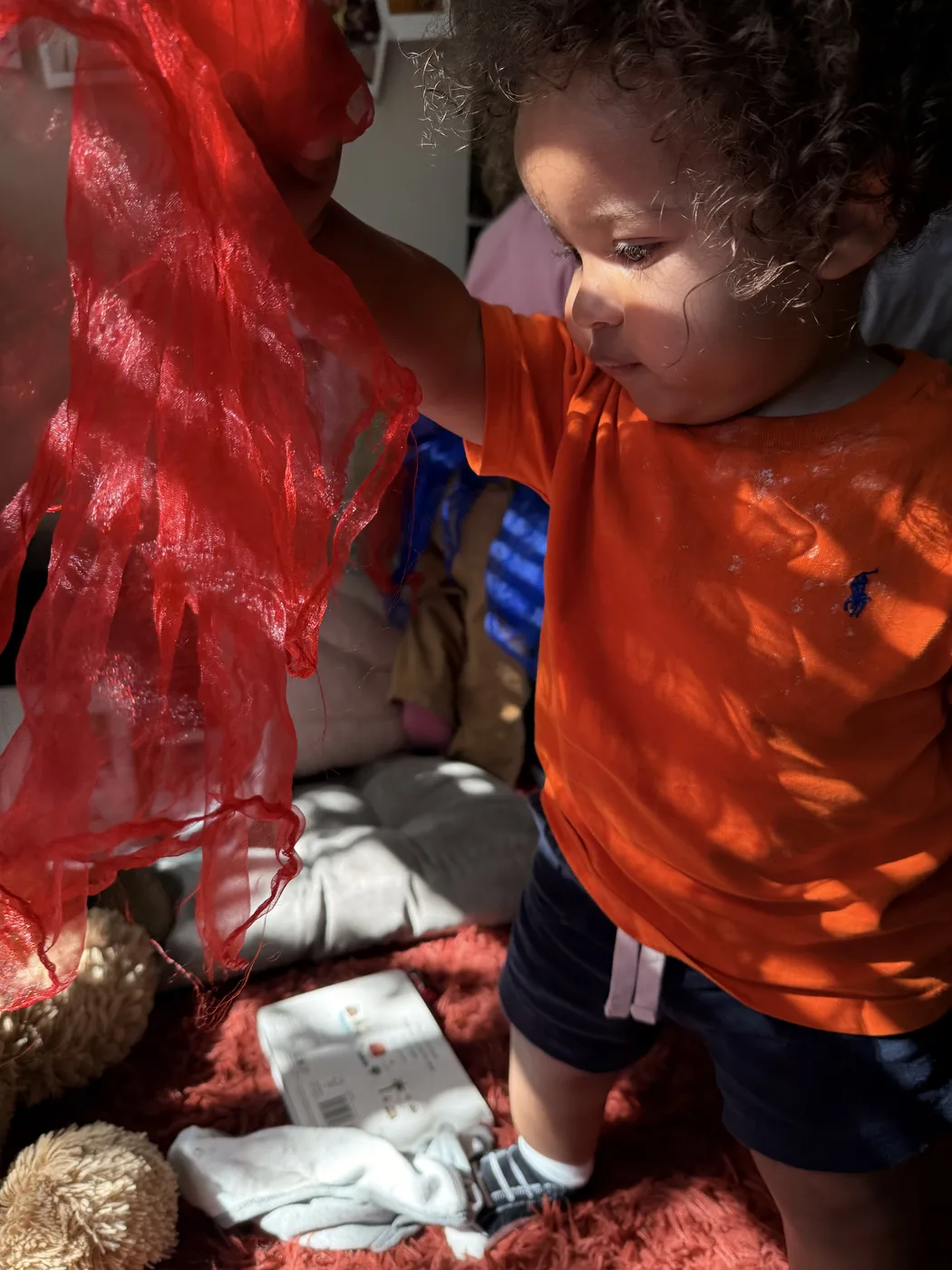 A toddler pulling a length of red sheer fabric across the play space