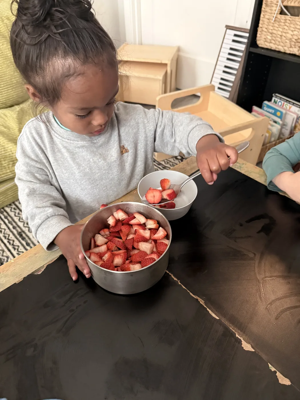 A child spooning cut strawberries into a metal bowl at a low table
